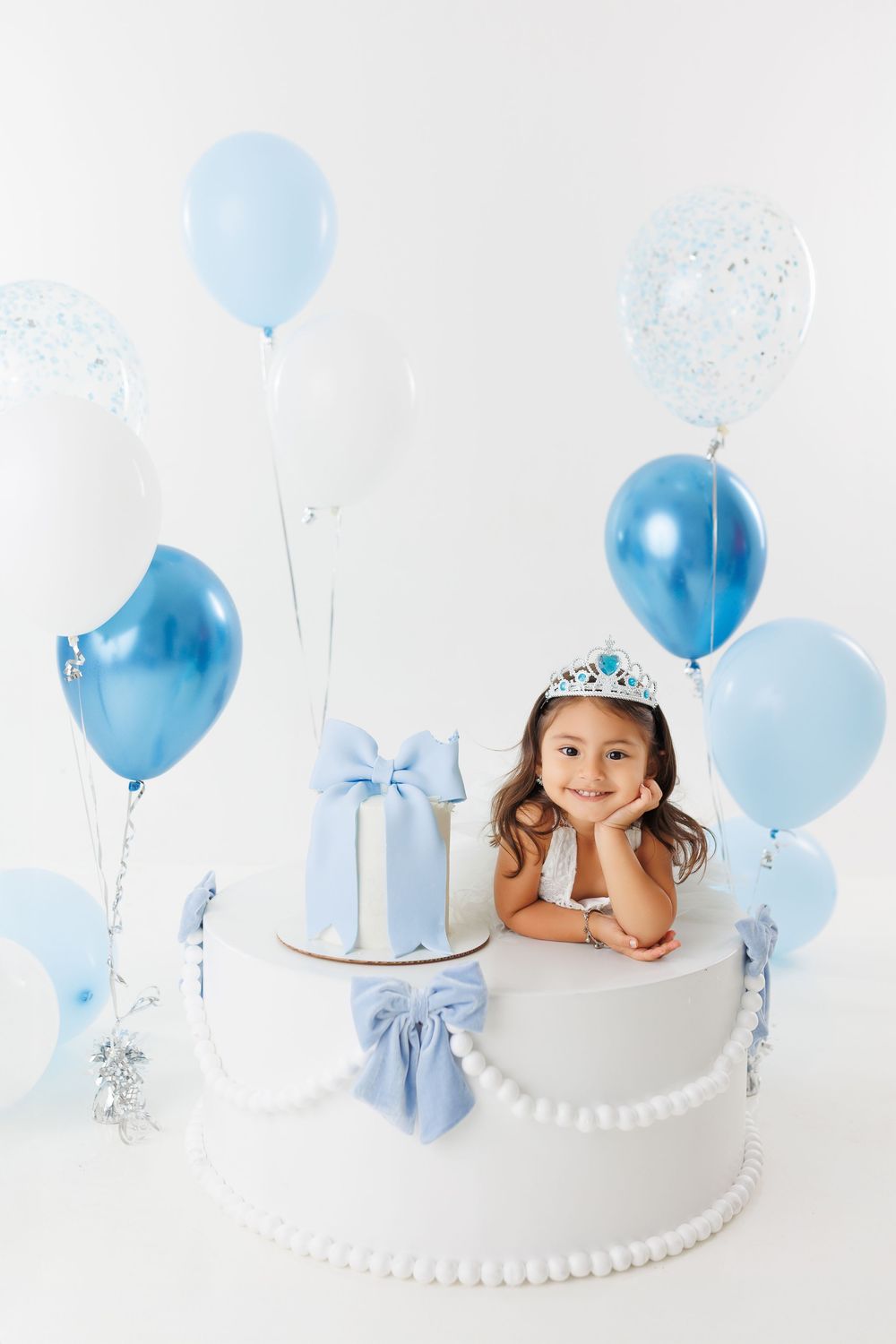 Cake smash photoshoot setup with blue balloons, white backdrop, and silver crown for baby's first birthday celebration.