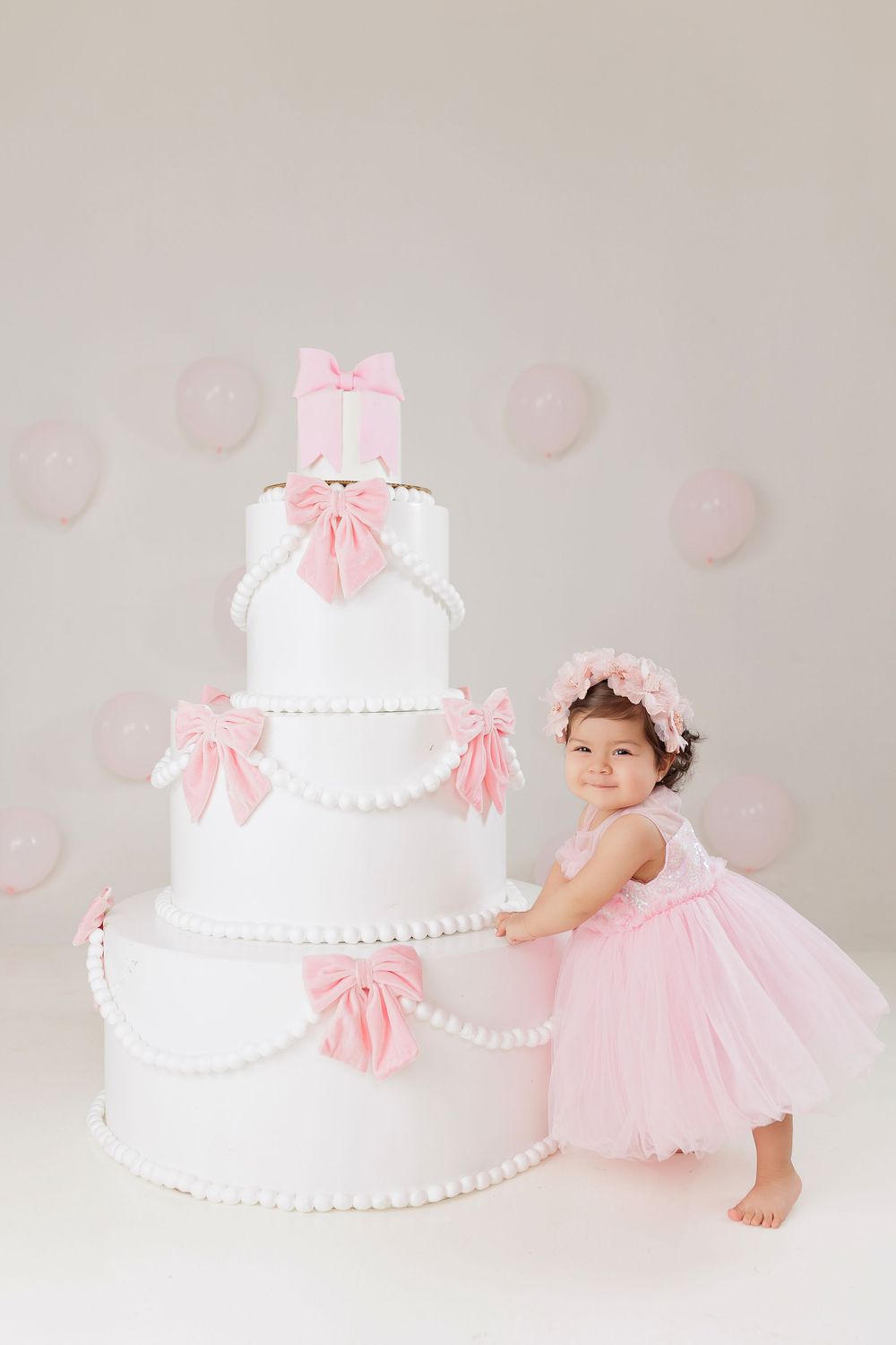 A large white tiered cake with pink bows sits beside a small child in a pink tulle dress and floral crown.