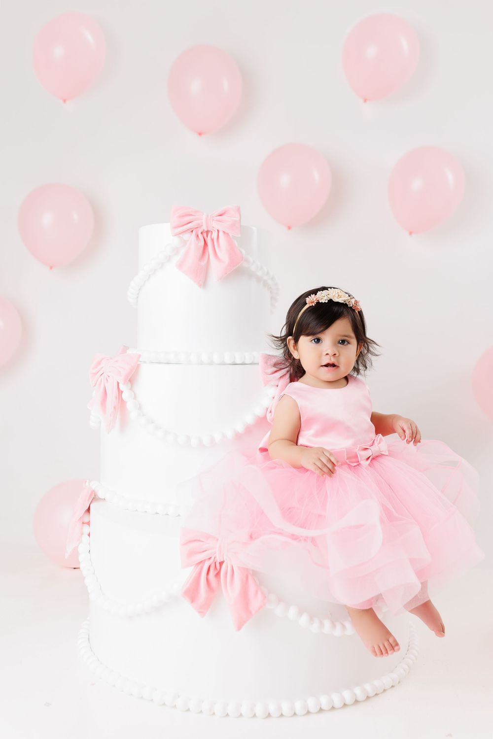 Baby in pink tulle dress poses against white backdrop with pink balloon decorations for first birthday photoshoot.