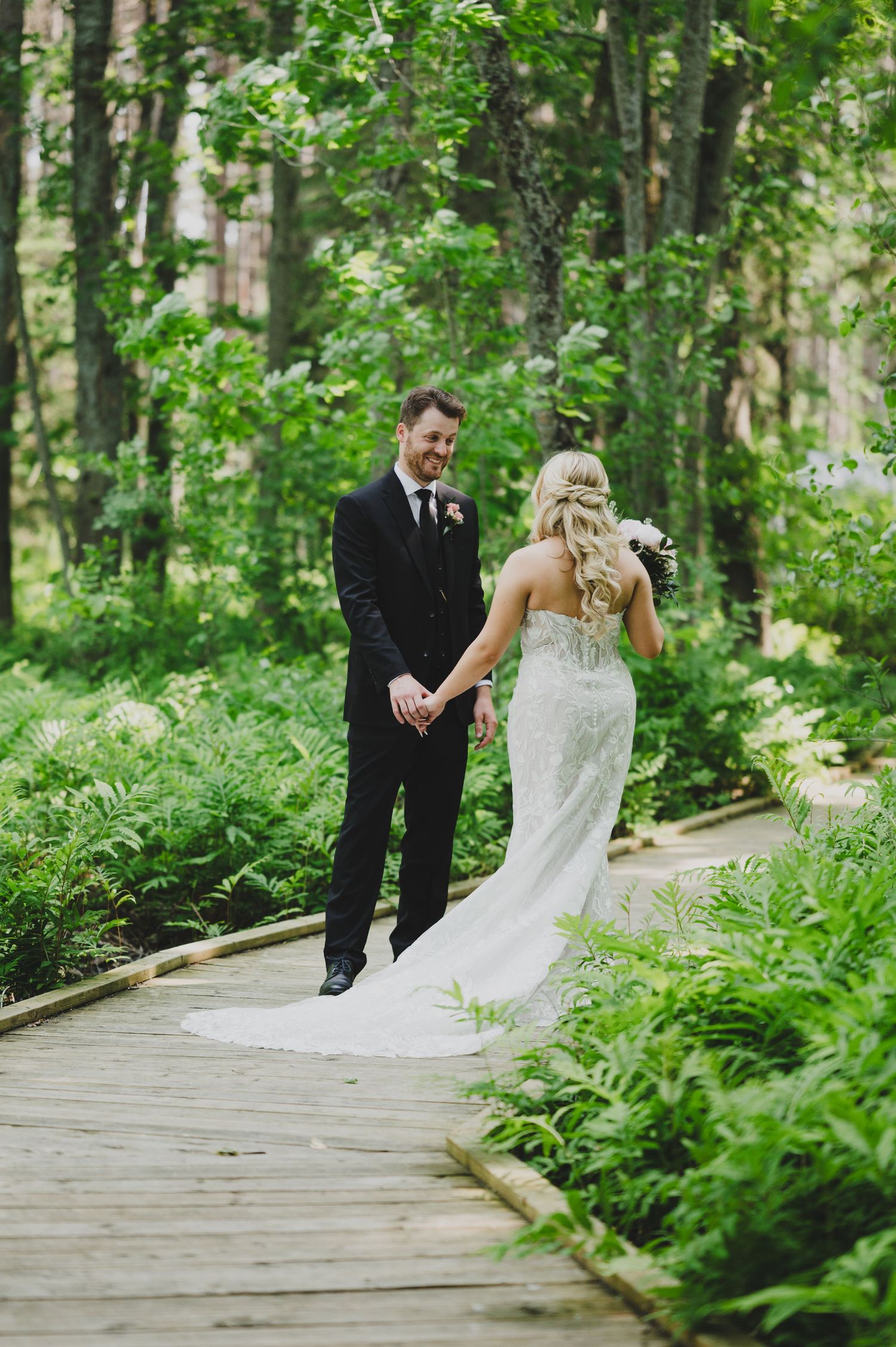 Newlyweds walk together on a wooden boardwalk surrounded by lush green forest ferns and trees.