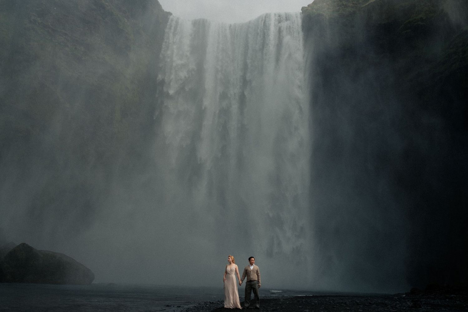 Couple standing together before massive Skogafoss waterfall in misty Iceland landscape.