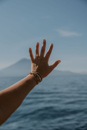 Hand reaching toward blue ocean horizon with mountains in background against clear sky.