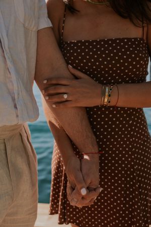 Close-up of couple holding hands by the ocean, wearing polka dot dress and beige pants in warm sunlight.