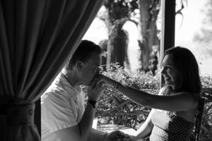 A romantic black and white moment between a couple sharing a sweet gesture by a curtained window.