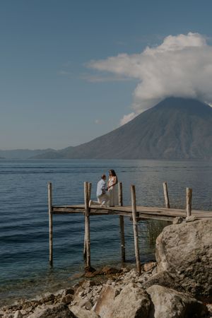 Couple on a rustic wooden dock with a volcanic mountain and blue lake backdrop.