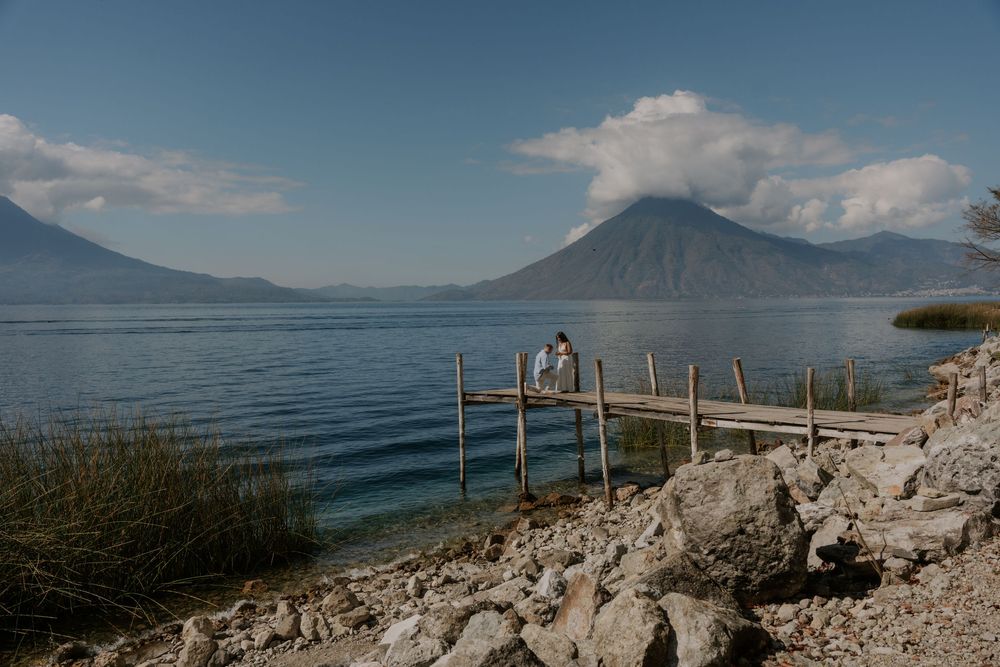 Couple standing on a wooden dock at Lake Atitlan with volcanic mountains in the background.
