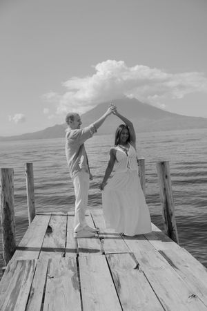 Black and white photo of a couple dancing on a pier with a volcano and ocean behind them.