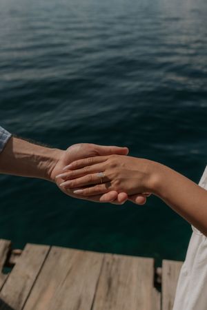 Couple holding hands on a wooden dock overlooking calm blue water, woman wearing an engagement ring.