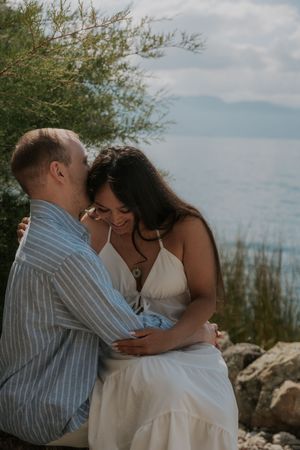 Romantic couple embracing on rocky shore with trees and calm blue water in the background.