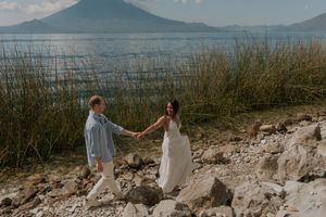 Couple holding hands on rocky shore with tall grass and volcanic mountain behind them.