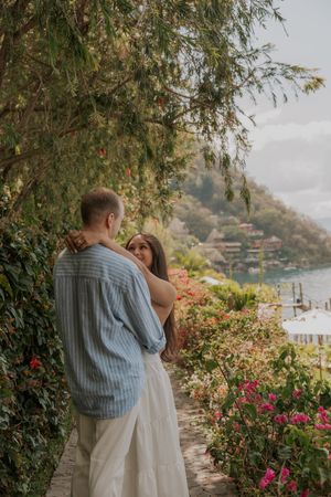 Romantic couple embracing on a lush garden path with colorful flowers and a scenic lake view.