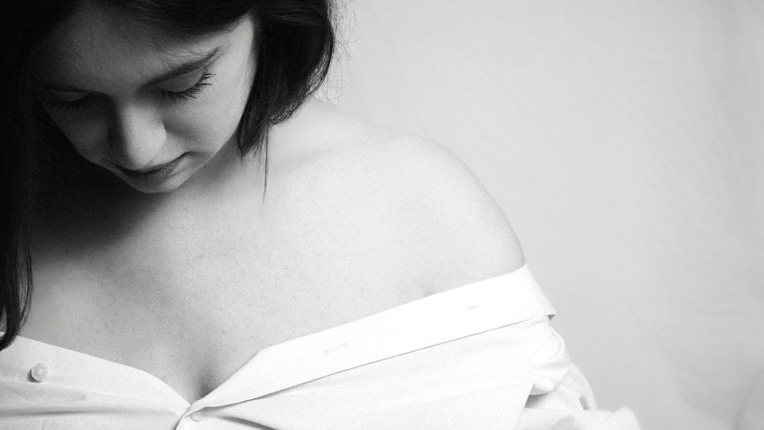 Boudoir style photoshoot with as woman looking down, she in in a white shirt that is showing her solders