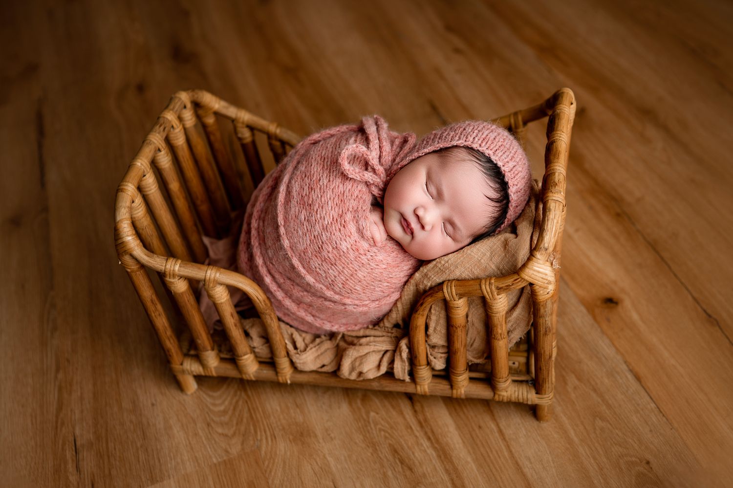 A sleeping newborn in a pink wrap lies peacefully in a wooden basket on hardwood flooring.