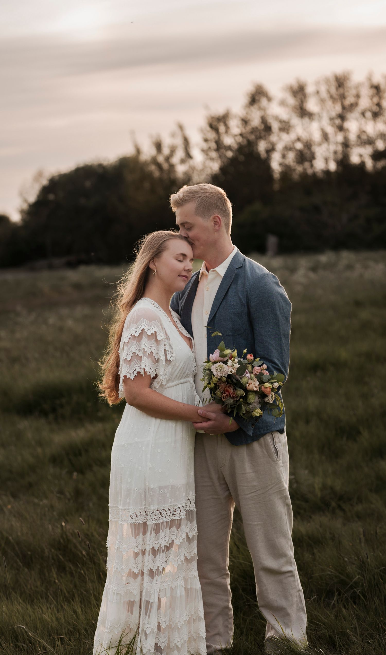 bröllopsfotografering på en äng. Bruden bär en vit lång klänning och håller en bukett. Brudgummen  en vit skjorta, blå kavaj och beiga byxor