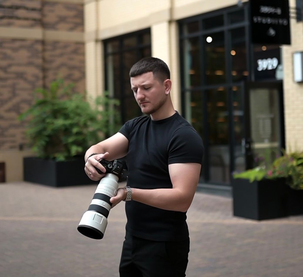 A person in a black t-shirt examines a camera lens while standing outside a brick building.