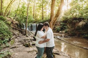 Couple sharing a romantic moment near a sunlit waterfall surrounded by lush green forest trees.