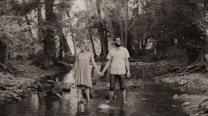 Couple holding hands standing in a shallow creek surrounded by lush trees in a vintage black and white photo.