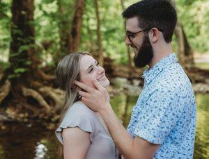 A smiling couple shares a tender moment outdoors near a creek, man gently touching woman's face.