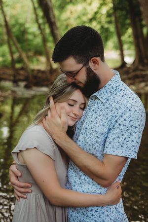 A couple embraces tenderly outdoors near a creek, man in floral shirt gently cupping woman's face as she smiles.