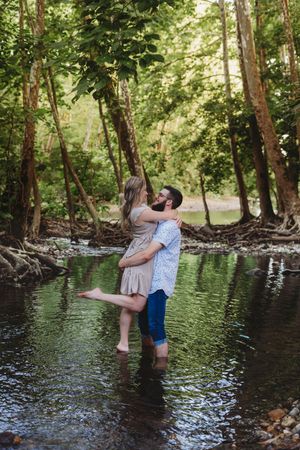 Couple embracing romantically in a shallow forest creek surrounded by lush green trees.