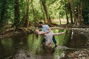 Couple romantically dancing in a shallow forest creek surrounded by lush green trees and exposed roots.