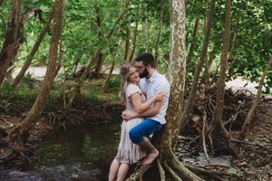 Couple embracing romantically on tree roots by a creek in a lush green forest setting.