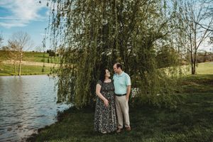 Couple standing under a weeping willow tree by a pond in a lush green park during golden hour.