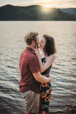 Couple sharing a romantic moment by a lake at sunset, woman in floral jumpsuit embracing man in red shirt.