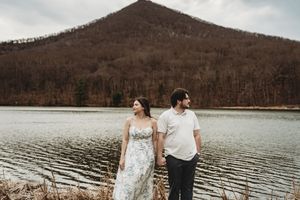 Couple holding hands by a serene lake with a forested mountain backdrop during an outdoor engagement session.