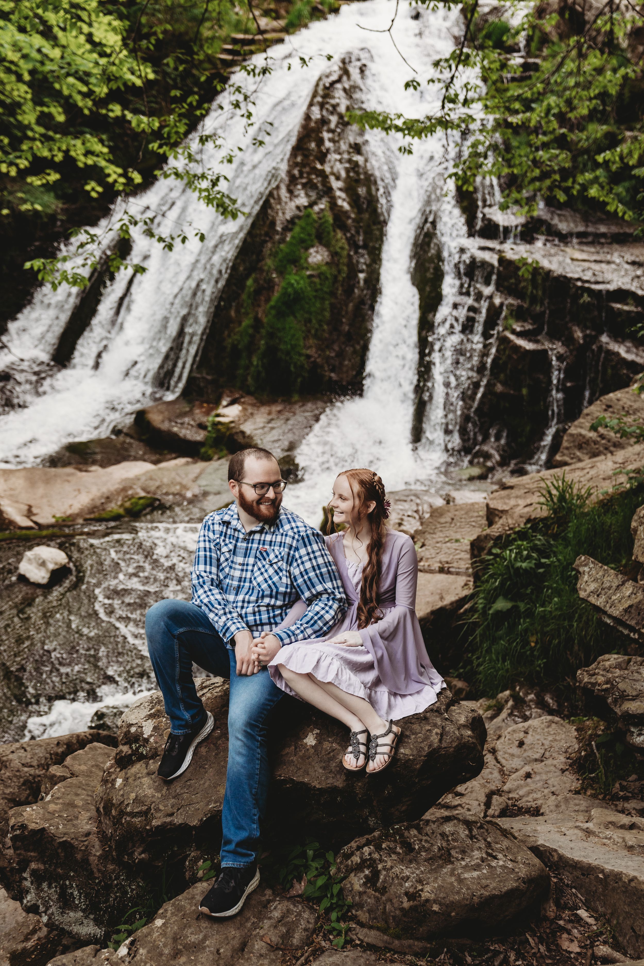 Couple sitting on rocks holding hands in front of a cascading waterfall surrounded by lush green trees.