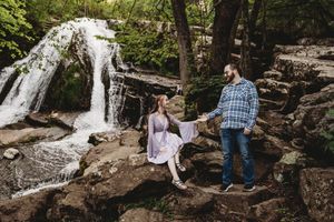 Couple holds hands on rocks near a waterfall surrounded by lush green trees in a romantic outdoor setting.