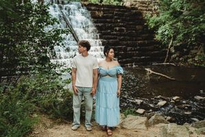 Couple holding hands in front of a stone waterfall surrounded by lush greenery, wearing casual summer outfits.