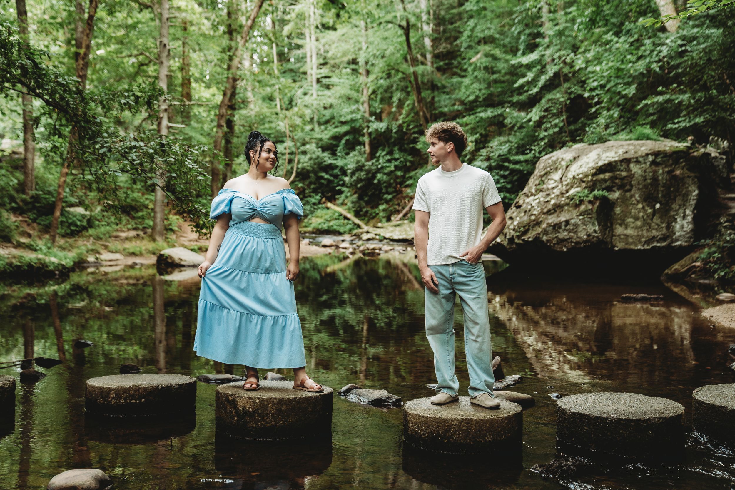 Couple standing on stepping stones in a lush green forest creek, woman in blue dress, man in white shirt.