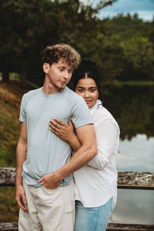 A young interracial couple embraces near a serene lake surrounded by lush green trees outdoors.