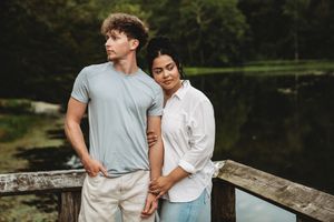 A young interracial couple poses on a wooden bridge near a serene lake surrounded by lush green trees.