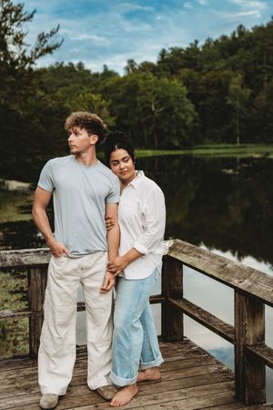 Couple standing on a wooden dock by a serene lake surrounded by lush green trees on a cloudy day.