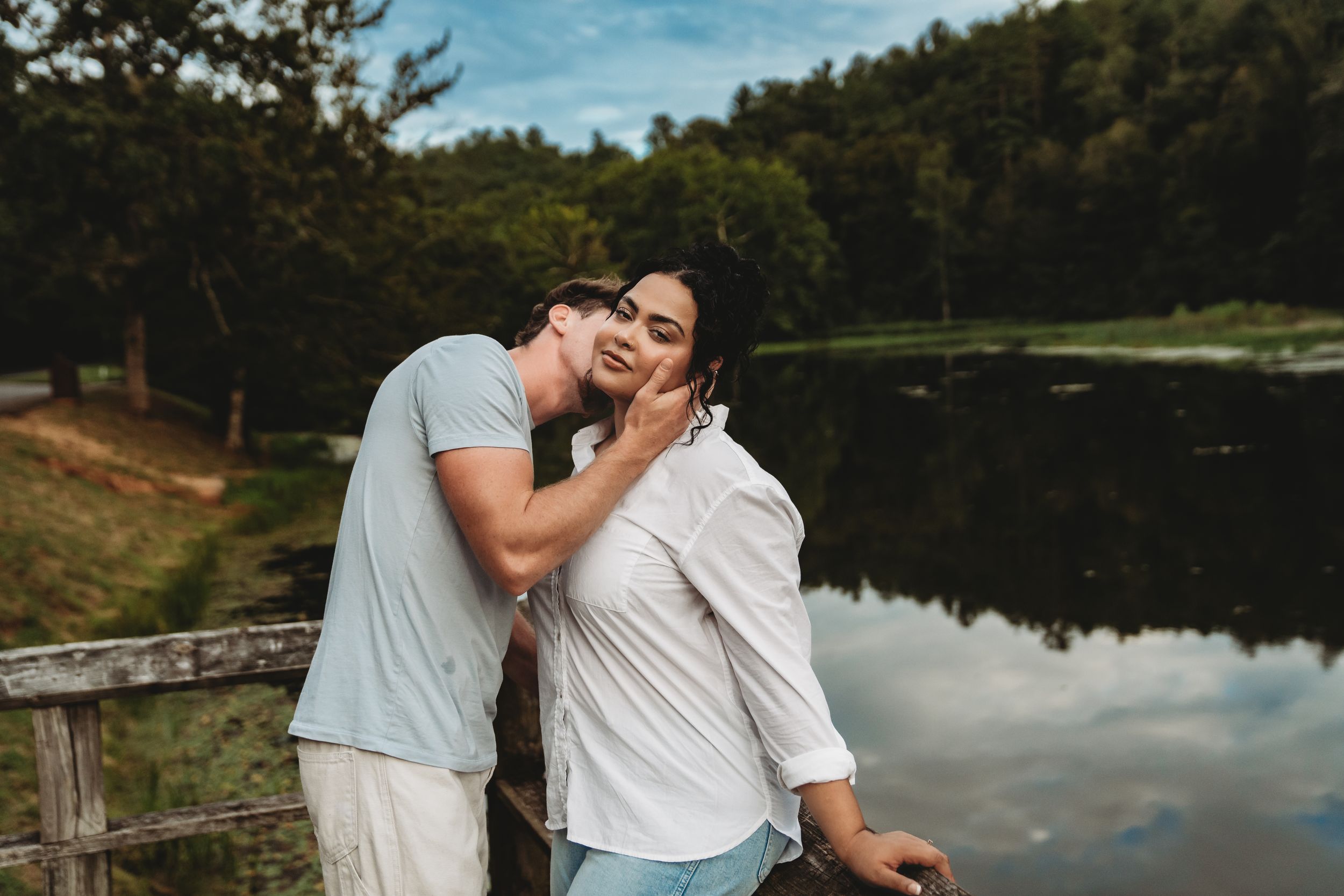 Romantic couple embracing by a serene lake surrounded by lush green trees during a warm summer day.