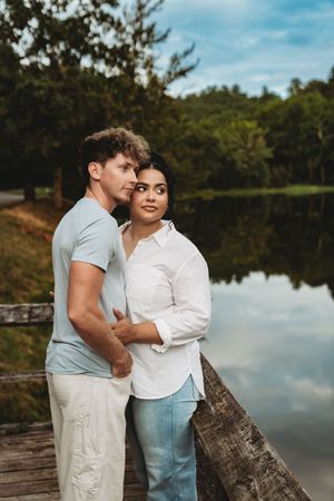 Couple embracing on a wooden dock by a serene lake surrounded by lush green trees under a cloudy sky.