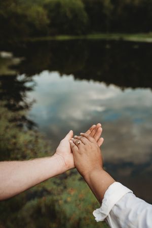 Two hands touching near a reflective lake, one wearing an engagement ring, symbolizing love and commitment outdoors.