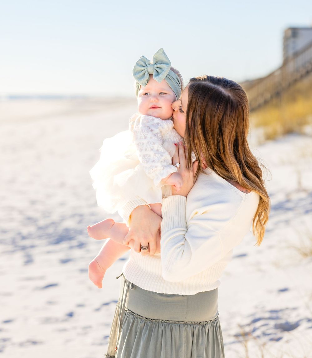 A parent and baby share a tender moment on a sunny beach with white sand and coastal dunes in the background.