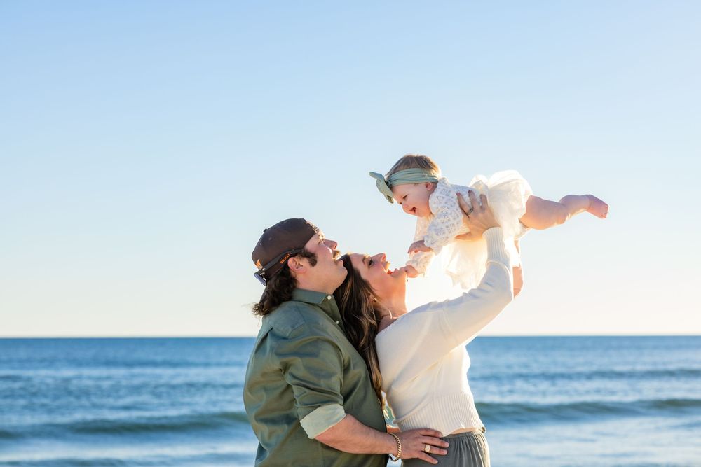 A person in a hat and jacket lifts a baby in the air against a blue ocean backdrop at sunset.