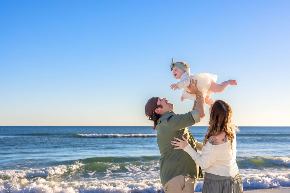 A family enjoys a playful moment together at the beach with crashing waves and blue sky in the background.