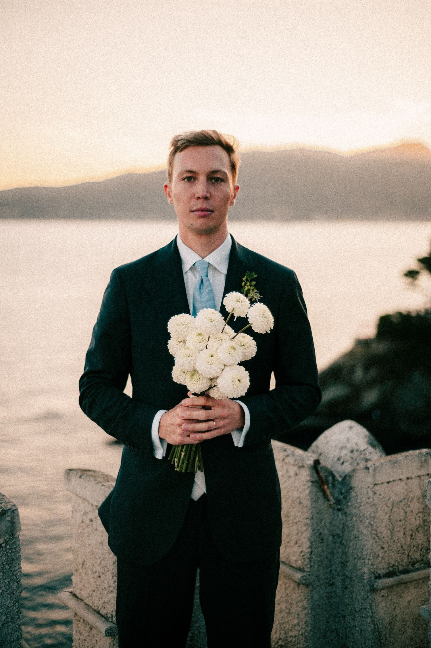 A groom in a black suit holds a white rose bouquet against a scenic waterfront backdrop at sunset.