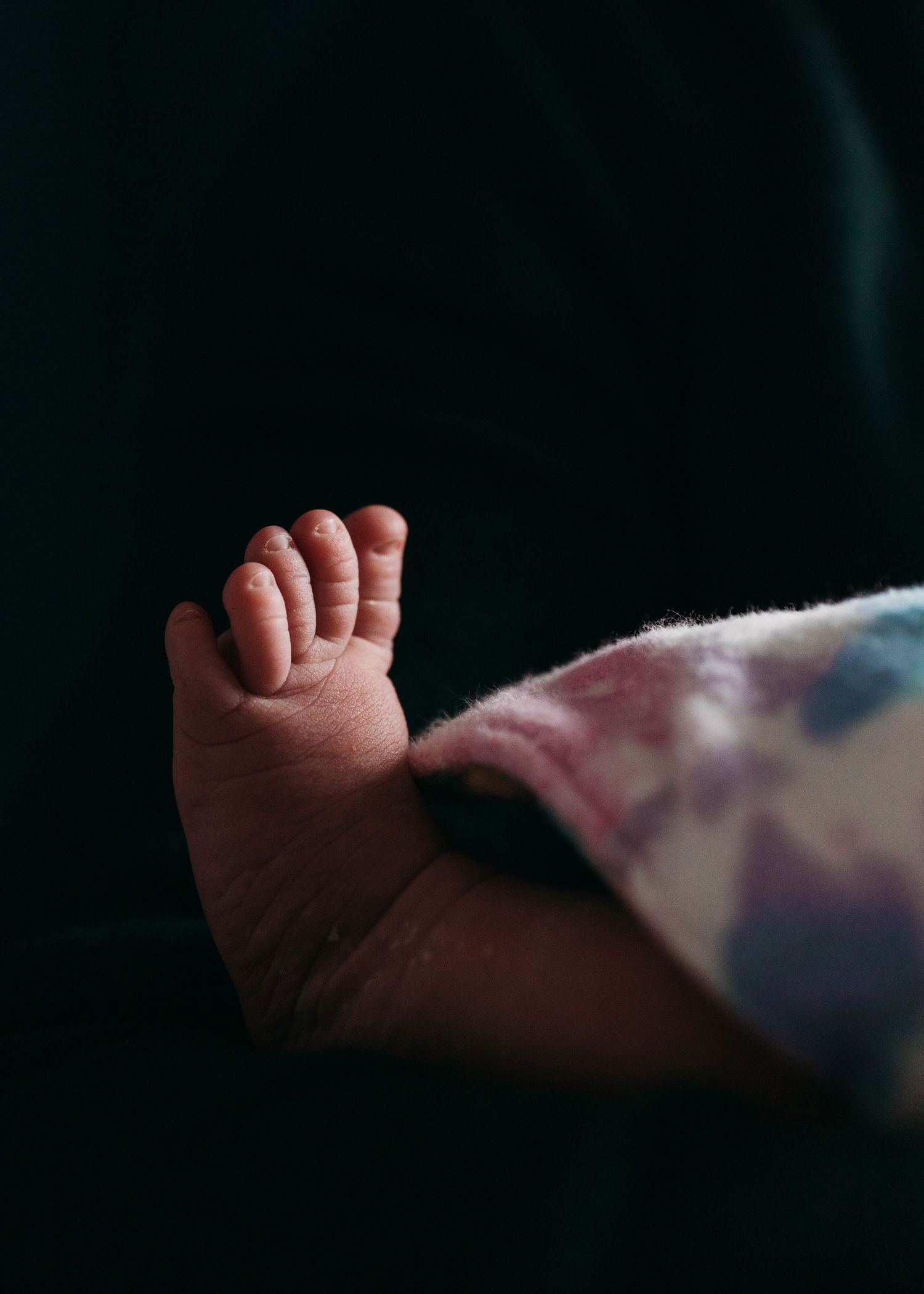 Close-up view of tiny newborn baby toes peeking out from under a floral blanket against dark background.
