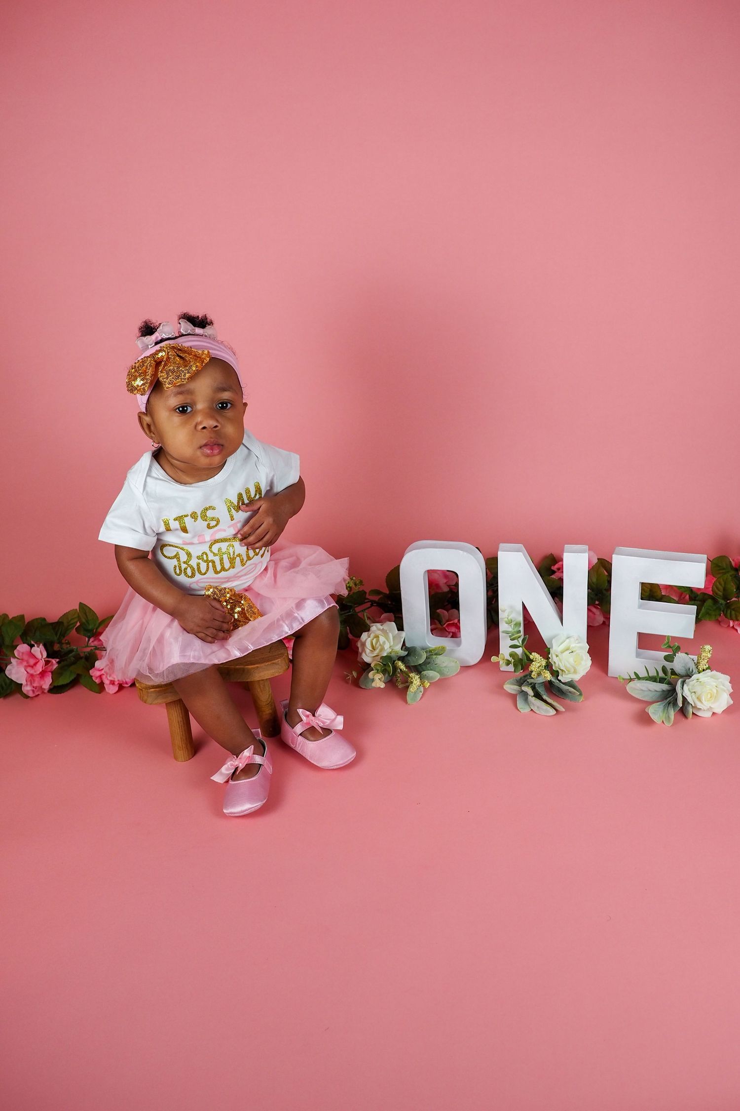 First birthday photo setup with decorative 'ONE' letters and floral accents on pink background.
