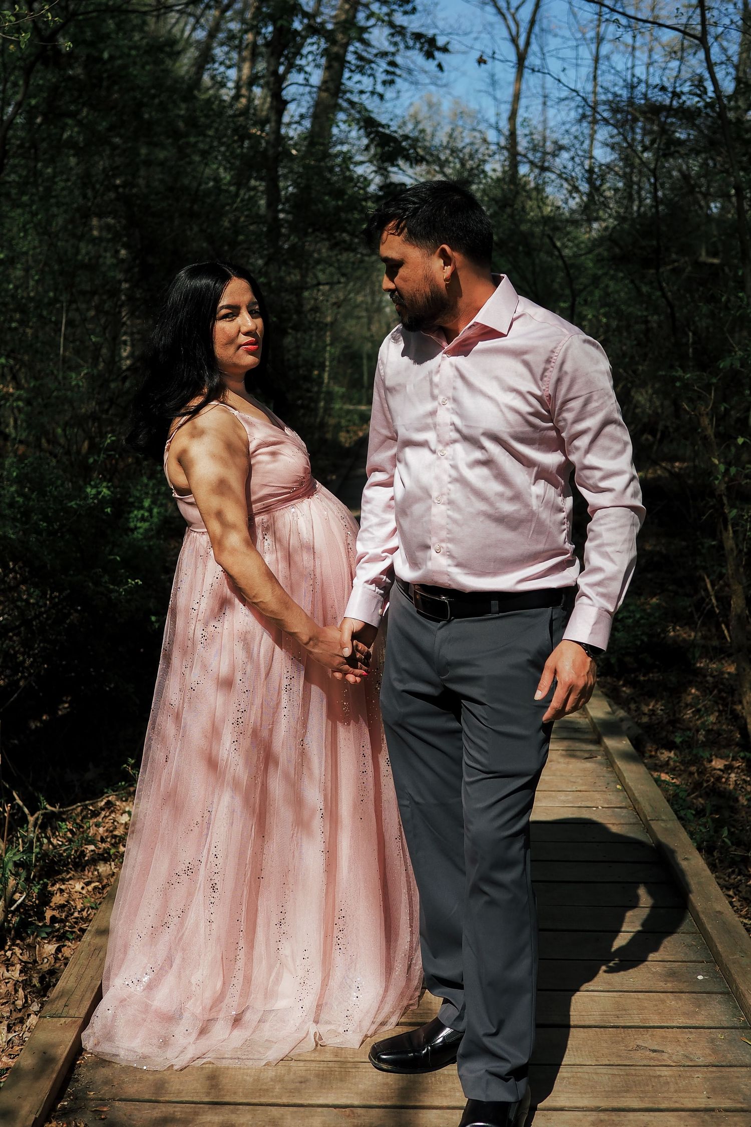 A romantic maternity photo of a couple on a wooden boardwalk in a forest setting.