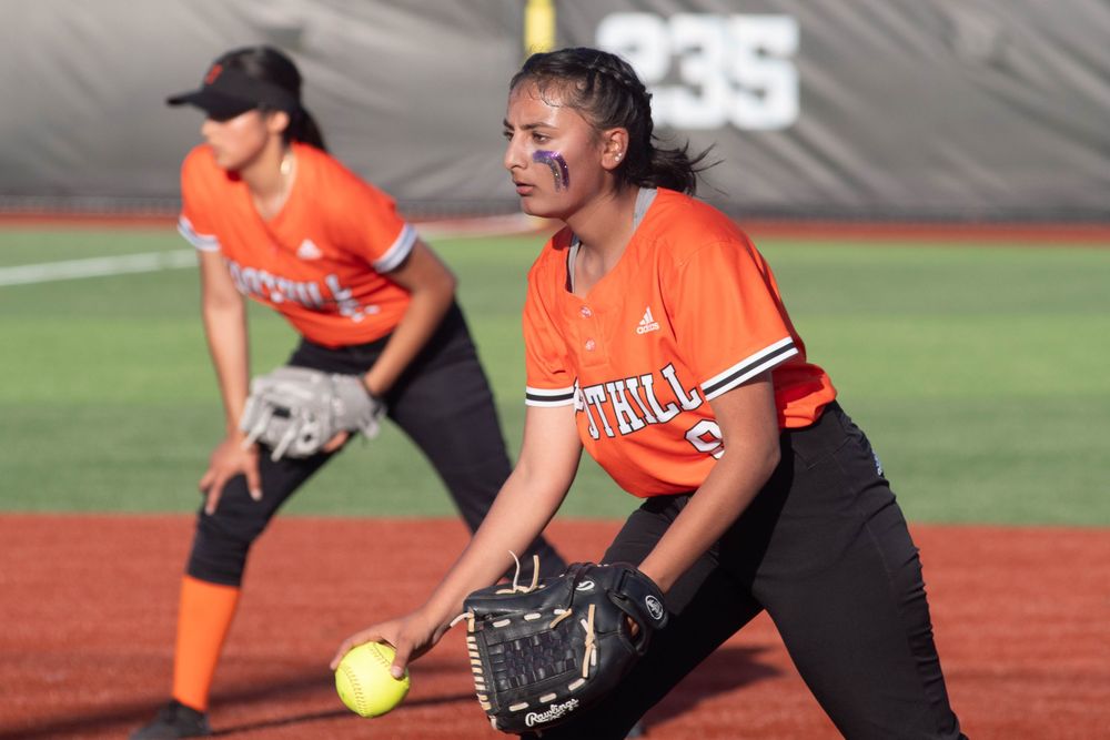 A softball player in an orange jersey pitches the ball during a game.