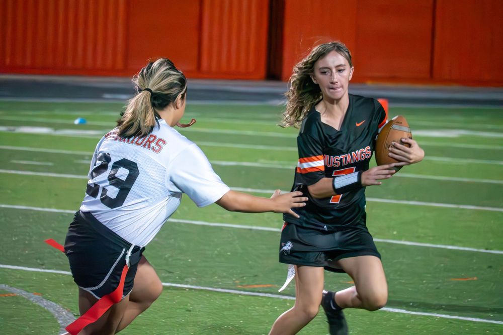 Female flag football players compete for the ball during an intense game on an artificial turf field.