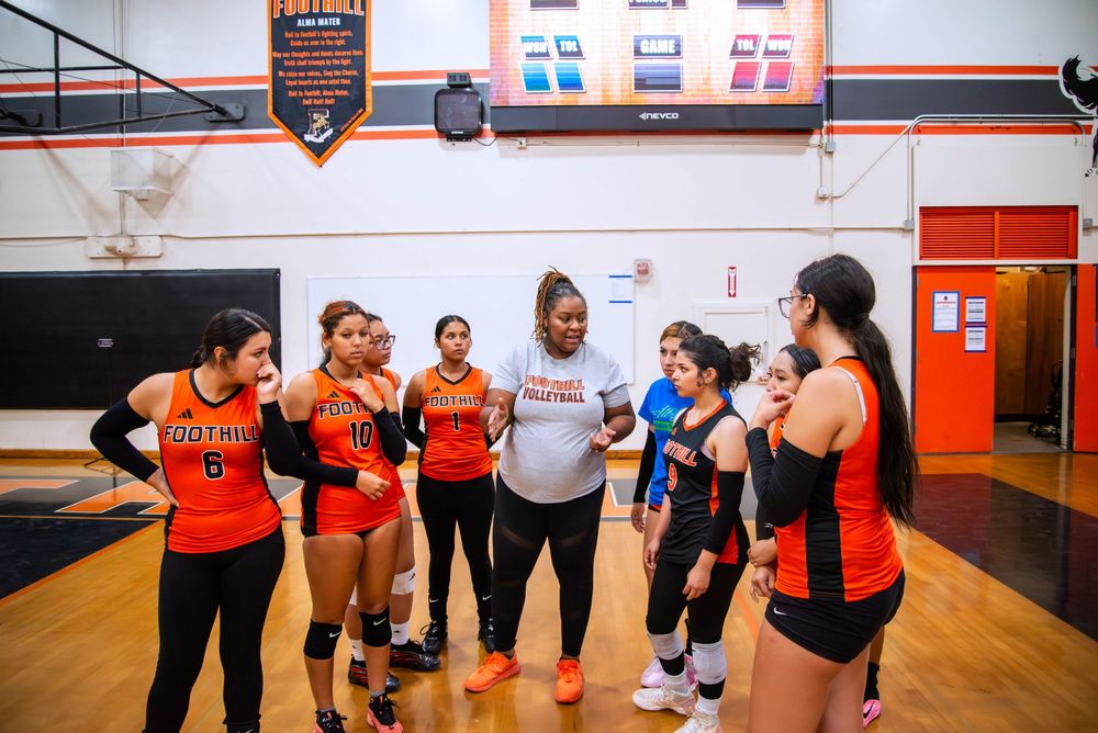 A group of athletes in orange and black uniforms gather around their coach during practice in a gymnasium.