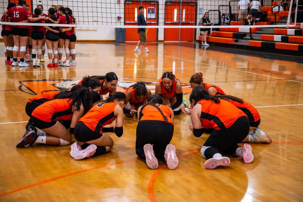 Volleyball team huddles together in orange uniforms on gymnasium court before match.